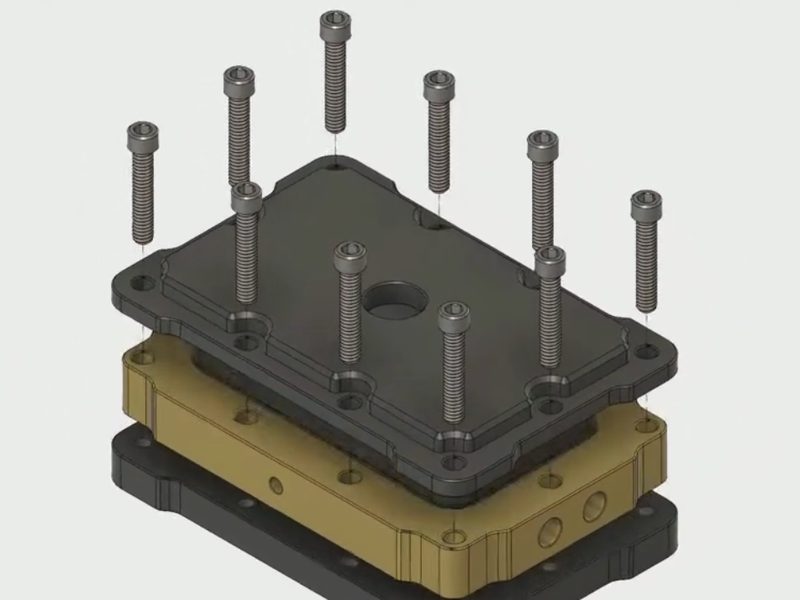 An exploded CAD view of a three-plate milled assembly: a black cover
                      with a bored central hole, a brass body between, and a black base,
                      with twelve socket head cap screws lifted out above their holes.