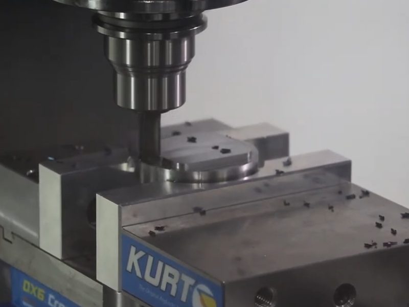 An end mill cutting a circular boss on a steel block held in a Kurt
                      vise on a CNC mill, with chips scattered across the vise and parallels.
