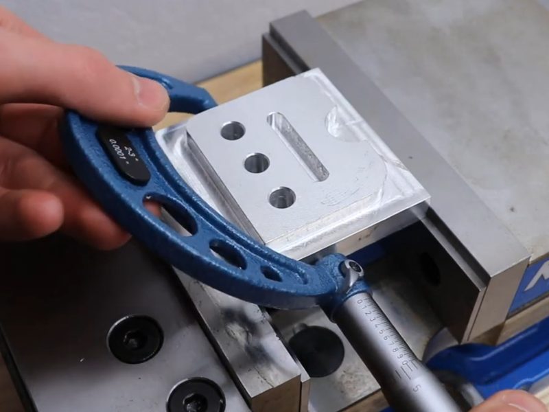 A two-to-three inch micrometer being closed onto a machined aluminium
                      plate in a vise. The plate carries three drilled holes and a milled
                      slot.