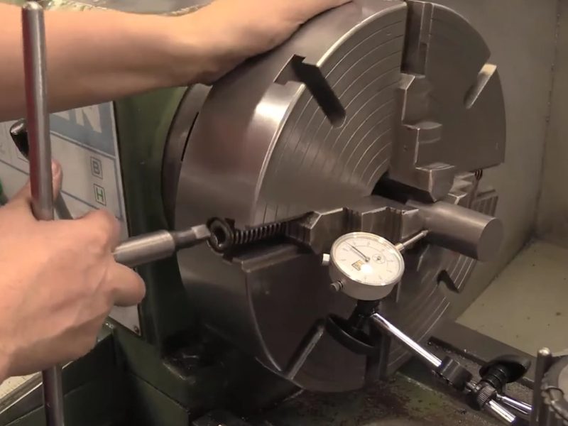 A student checking runout on a bar held in a four-jaw lathe chuck,
                      turning the chuck by hand with a key while a dial indicator rests
                      against the bar.