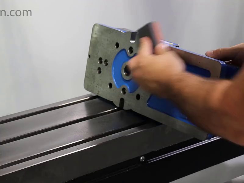 A machinist lowering a milling vise onto the T-slotted
                        table of a machining centre, both hands under it, the
                        fixed jaw facing across the slots.